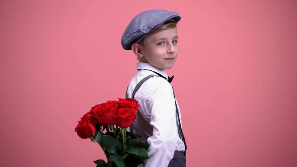 Little gentleman kid hiding roses behind back and turning to camera, smiling - Photo, Image