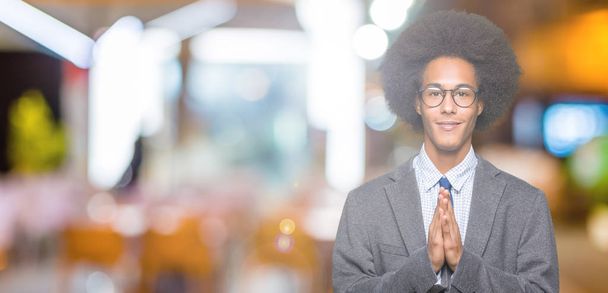 Young african american business man with afro hair wearing glasses praying with hands together asking for forgiveness smiling confident. - Photo, Image