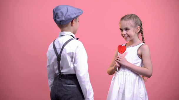 Little boy gifting red heart-shaped lollipop to girlfriend, sweet romantic gift - Fotó, kép