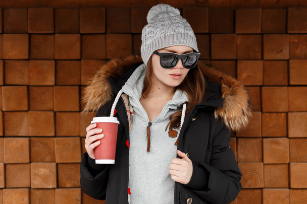 Çekici genç yenilikçi bir ahşap duvar poz ve bir fincan kahve elinde tutan güneş gözlükleri şık bir ceket bir kazak örme şapka kadında. Güzel kız kış günü - Fotoğraf, Görsel