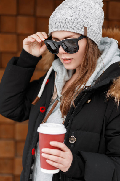 Elegante giovane hipster donna in cappello a maglia con una felpa blu in una giacca invernale in occhiali da sole in piedi vicino alla parete di legno e in possesso di una tazza rossa di caffè caldo gustoso. Ragazza alla moda
. - Foto, immagini