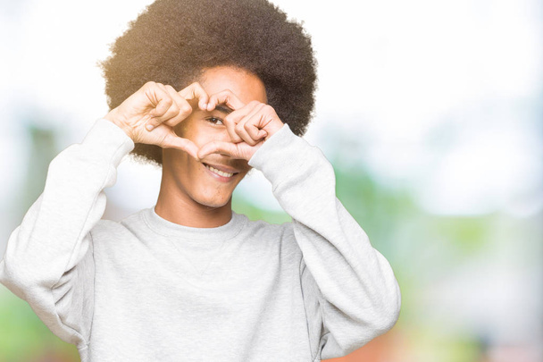 Young african american man with afro hair wearing sporty sweatshirt Doing heart shape with hand and fingers smiling looking through sign - Fotografie, Obrázek