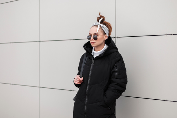 Modern young hipster woman in round sunglasses in a stylish coat in a fashionable bandana with a leather backpack is standing near a white wall a warm winter day. Fashionable american girl on vacation - Fotó, kép