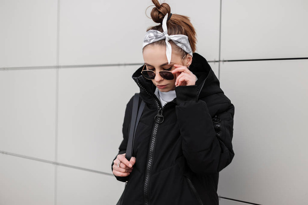 Mujer hipster joven moderna con un peinado elegante en un pañuelo en una chaqueta negra de moda con una mochila elegante en un golf de punto con gafas de sol elegantes posando cerca de un edificio blanco. Chica de moda
. - Foto, imagen
