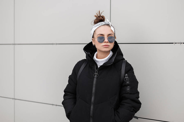 Европейская стильная молодая женщина-хипстер в модном черном пальто с модной прической со стильной банданой, позирующей в городе возле белой стены. Женская одежда в американском стиле
. - Фото, изображение
