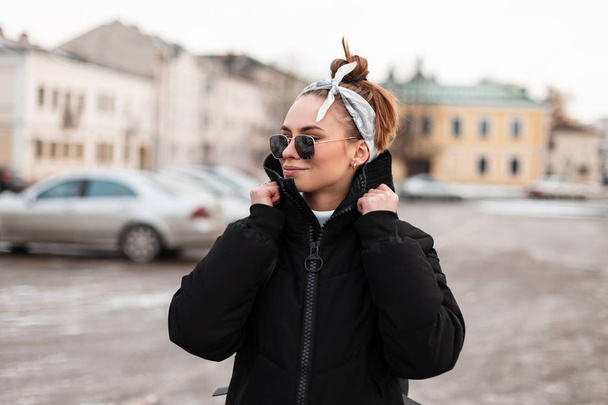 Mujer hipster joven atractiva moderna en ropa elegante de invierno con un peinado con un pañuelo en gafas de sol en la ciudad en el fondo de los edificios antiguos. Elegante chica americana viaja
. - Foto, Imagen