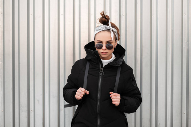 junge modische schöne Hipster-Frau mit stylischer Frisur und Kopftuch in schwarzer Sonnenbrille in einem langen schwarzen Mantel mit einem Rucksack auf den Schultern posiert in der Nähe einer Metallwand. modernes stilvolles Mädchen - Foto, Bild