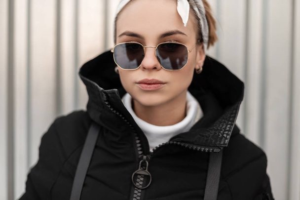 Retrato de uma jovem mulher hipster na moda em óculos de sol da moda com uma bandana no golfe branco em uma jaqueta elegante com uma mochila no fundo de uma parede de metal. Menina moderna
.  - Foto, Imagem