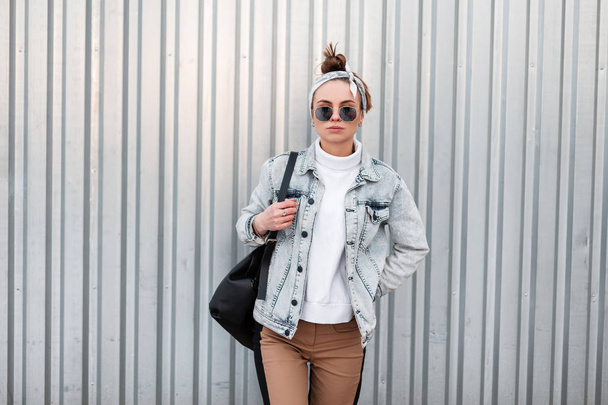 American hipster young woman with a stylish hairstyle in summer denim jackets in trendy pants in a knitted white sweater in sunglasses with a bandana near a shiny gray metal wall. Modern girl. - Photo, Image