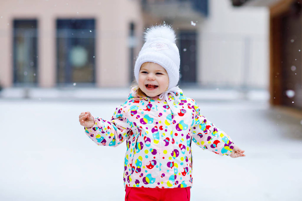 冬に屋外を歩く小さな幼児の少女の肖像画。かわいい幼児は甘いロリポップキャンディーを食べています。寒い雪の日に楽しんでいる子供。暖かい赤ちゃんのカラフルな服やボブル付きの帽子を身に着けて. - 写真・画像