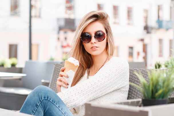 Charming attractive young hipster woman in a vintage stylish sweater in blue fashionable jeans in trendy sunglasses with ice cream in her hands is sitting in a street cafe. Cute girl. - Fotoğraf, Görsel