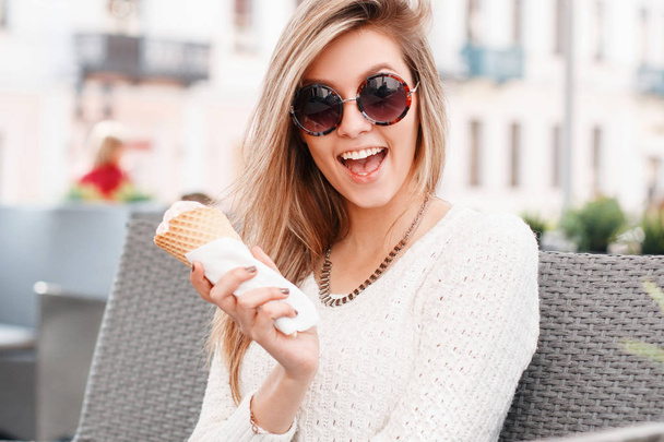 Funny cheerful young hipster woman in a white knitted vintage sweater with sweet vanilla ice cream in a cone sitting outdoors. Attractive joyful girl. - Foto, afbeelding