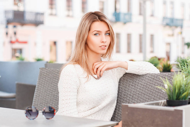Cute attractive young woman with blue eyes in a stylish knitted sweater is sitting on a vintage sofa in an outdoor cafe. Beautiful girl resting. - Foto, immagini