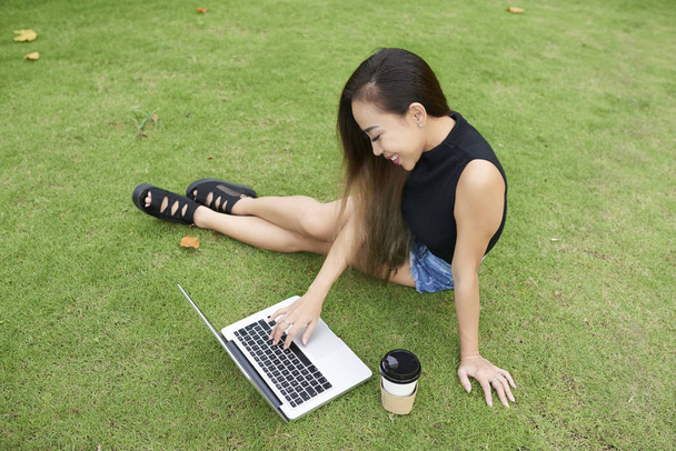 Cheerful young Vietnamese woman sitting outdoors and browsing laptop - Photo, Image