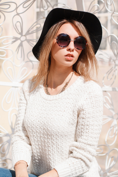 Πανέμορφο hipster νεαρή γυναίκα σε μοντέρνα γυαλιά ηλίου με λευκό πλεκτό πουλόβερ σε μπλε τζιν με ένα κομψό καπέλο κάθεται και ακουμπά σε ένα vintage κρέμεται μεταλλικό λευκό καρέκλα. Γοητευτικό κορίτσι. - Φωτογραφία, εικόνα