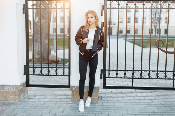 Όμορφη νεαρή γυναίκα με ένα κομψό σακάκι μαύρο με ένα άσπρο πουκάμισο πόλο σε μοντέρνα τζιν σε πάνινα παπούτσια λευκό στέκεται κοντά στις μεταλλικές vintage πύλες με φόντο το ηλιοβασίλεμα. Ελκυστική κοπέλα. - Φωτογραφία, εικόνα
