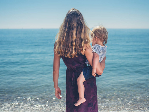 A young mother and her toddler child are by the water on the beach in summer - Foto, afbeelding