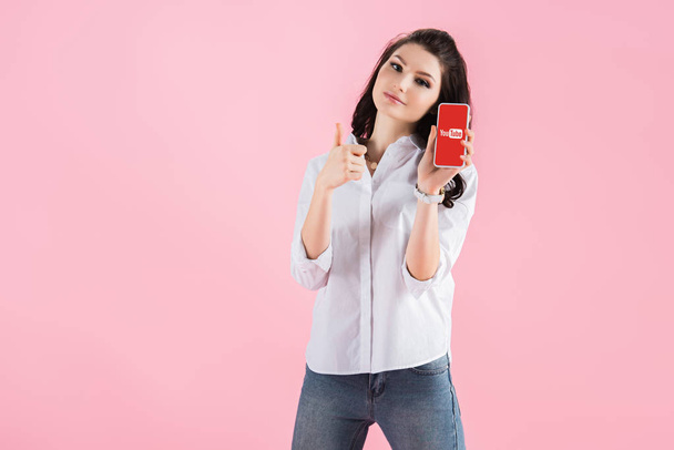 brunette girl showing thumb up and smartphone with youtube app on screen, isolated on pink  - 写真・画像