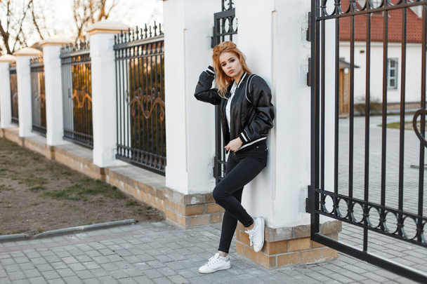 Stijlvolle jonge vrouw in een zwarte voorjaar jas in trendy jeans in een witte polo shirt in sneakers poseren in de buurt van een vintage metalen hek. Amerikaanse stijlvolle meisje. mode - Foto, afbeelding