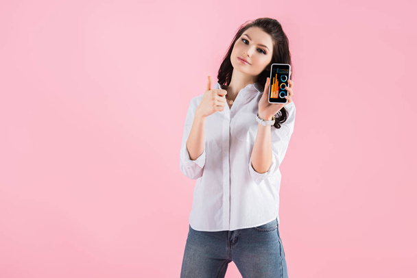 beautiful girl showing thumb up and smartphone with graphs on screen, isolated on pink - Photo, Image