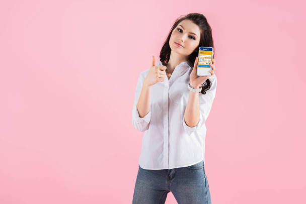 attractive brunette girl showing thumb up and smartphone with booking app on screen, isolated on pink  - Foto, Bild