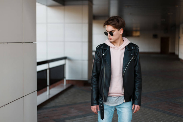Modne hipster młody człowiek w czarne okulary przeciwsłoneczne w Stylowa czarna kurtka w różowa Bluza w modny jeans w szary t-shirt stoi w pokoju. Poważne fajny facet z piękną fryzurę - Zdjęcie, obraz