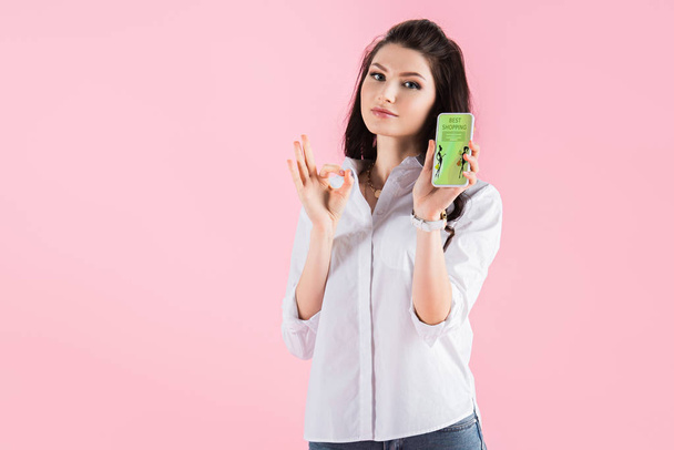 attraktive brünette Mädchen zeigt ok Zeichen und Smartphone mit Shopping-App auf dem Bildschirm, isoliert auf rosa - Foto, Bild