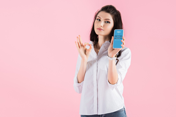 brunette girl showing ok sign and smartphone with skype app on screen, isolated on pink - Photo, Image