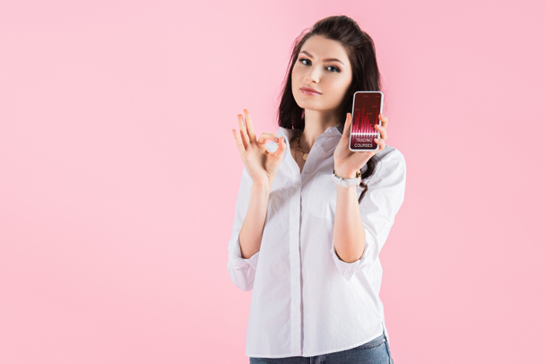 attraktive brünette Mädchen zeigt ok Zeichen und Smartphone mit Handelskursen auf dem Bildschirm, isoliert auf rosa - Foto, Bild