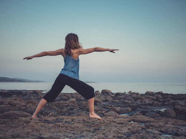 A young woman is doing yoga on the coast at sunset and is standing in a warrior pose - Foto, Imagen