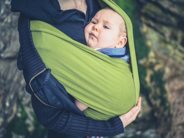 Uma jovem mãe está de pé na natureza com seu bebê em uma funda
 - Foto, Imagem
