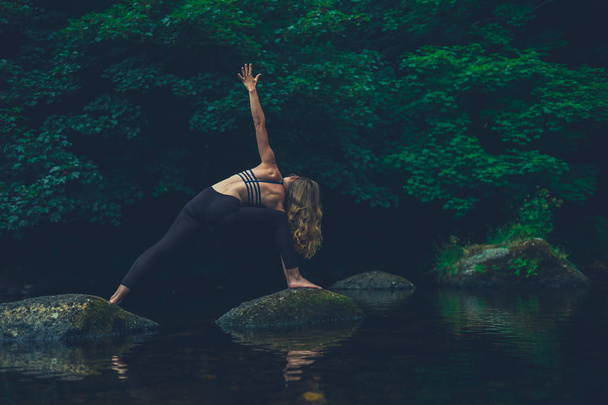A young woman doing yoga on a rock in the river is in a warrior pose - Photo, Image