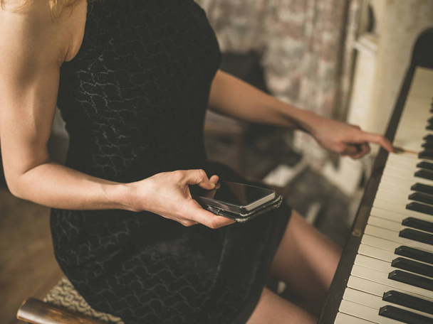 A young woman in a sexy dress is sitting at a piano and is using a smart phone - Фото, зображення