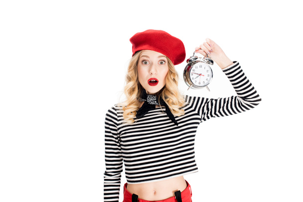 surprised woman in red beret holding alarm clock isolated on white  - Foto, afbeelding