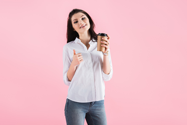 attraktives brünettes Mädchen mit Kaffee to go und Daumen hoch, isoliert auf rosa - Foto, Bild