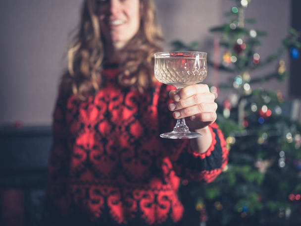 A happy young woman is standing by the christmas tree with a drink in her hand - Фото, изображение