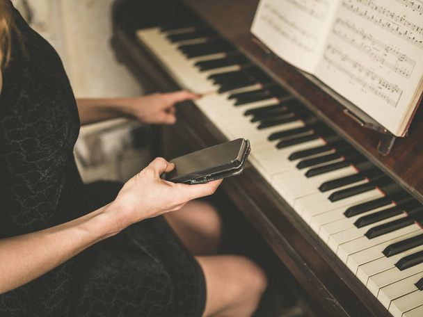 A young woman in a sexy dress is sitting at a piano and is using a smart phone - Foto, afbeelding
