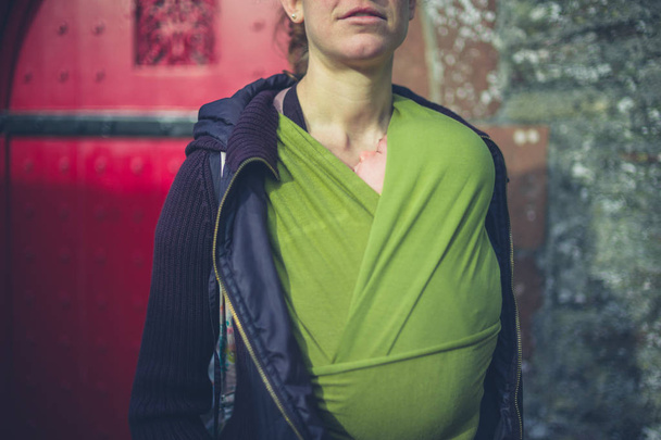 A young mother is standing by a red door and is carrying her baby in a sling - Foto, immagini