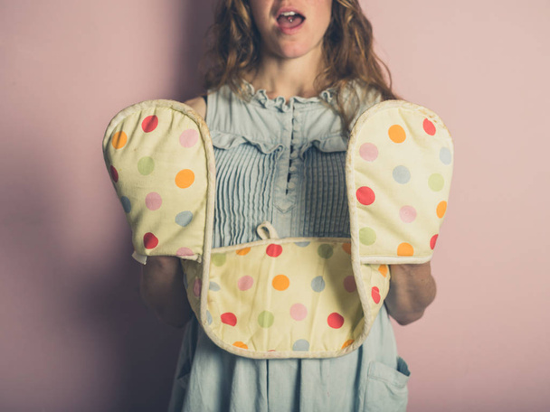 A surprised young woman is posing with oven gloves against pink background - Фото, изображение