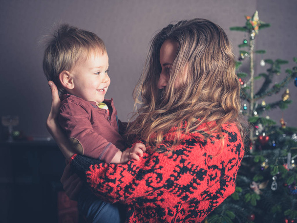 A young mother is standing by the christmas tree holding her smiling toddler - Photo, Image