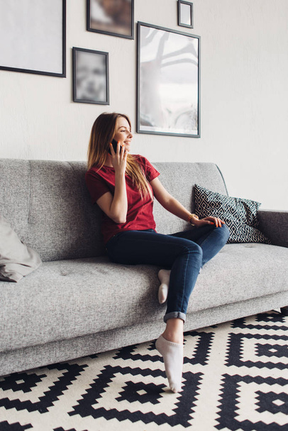 Молодая счастливая женщина сидит на диване и использует мобильный телефон дома
 - Фото, изображение