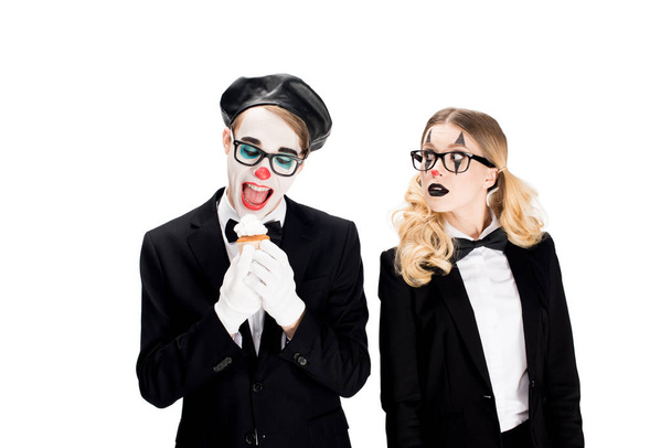 веселый клоун в костюме и очках, смотрящий на вкусный кекс рядом с женщиной, изолированной на белом
  - Фото, изображение