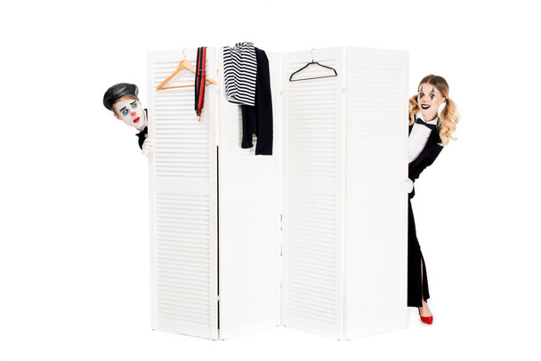 Lachende vrouw in pak kijken uit de kleedkamer deur in de buurt van verrast clown geïsoleerd op wit  - Foto, afbeelding