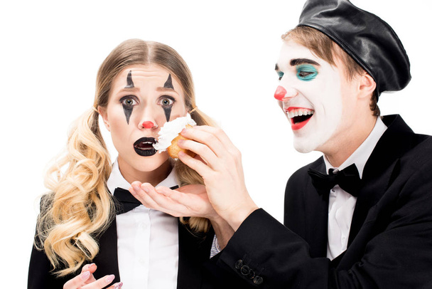 fröhlicher Clown wirft Cupcake ins Gesicht der überraschten Frau isoliert auf Weiß  - Foto, Bild