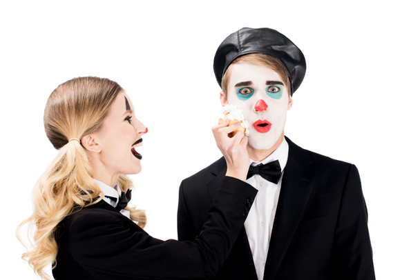 vrolijke vrouwelijke clown gooien cupcake ten opzichte van verrast man geïsoleerd op wit  - Foto, afbeelding