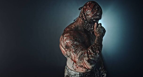 portrait of muscular zombie killer with mask covered in blood posing against misty background - Zdjęcie, obraz
