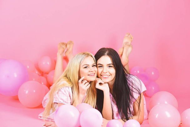 Blonde and brunette on smiling faces have fun at domestic party. Girls lay on belly near balloons, pink background. Friendship concept. Sisters or best friends in pajamas at girlish pajamas party - Foto, immagini