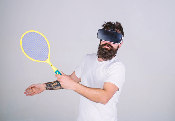 Guy with VR glasses play tennis with racket and ball. Man with beard in VR glasses beating pitch, grey background. Virtual sport concept. Hipster on busy face use modern technology for sport games - Foto, Bild