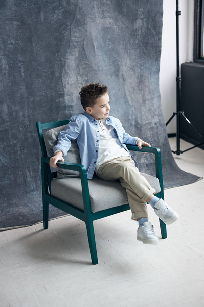Fashionable boy in blue shirt  sitting on chair in loft studio, grey backgroung. - Zdjęcie, obraz