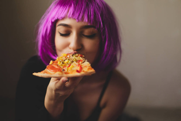Смешной розовый парик девушка в черном нижнем белье и рубашка ест пиццу дома. Девушка наслаждается вкусной пиццей
.  - Фото, изображение